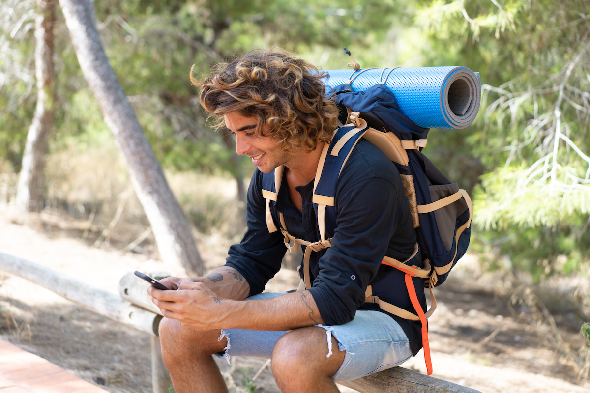 Male camper in the woods with a hiking backpack on sitting on a log while he is using his cell phone. 